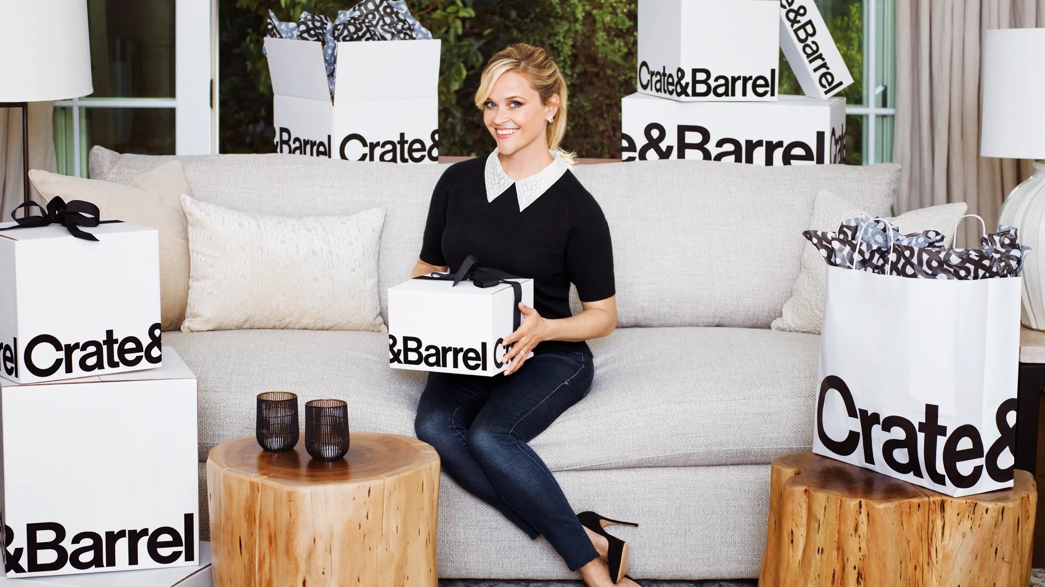 Reese witherspoon sitting on a sofa holding a white box labeled crate and barrel.